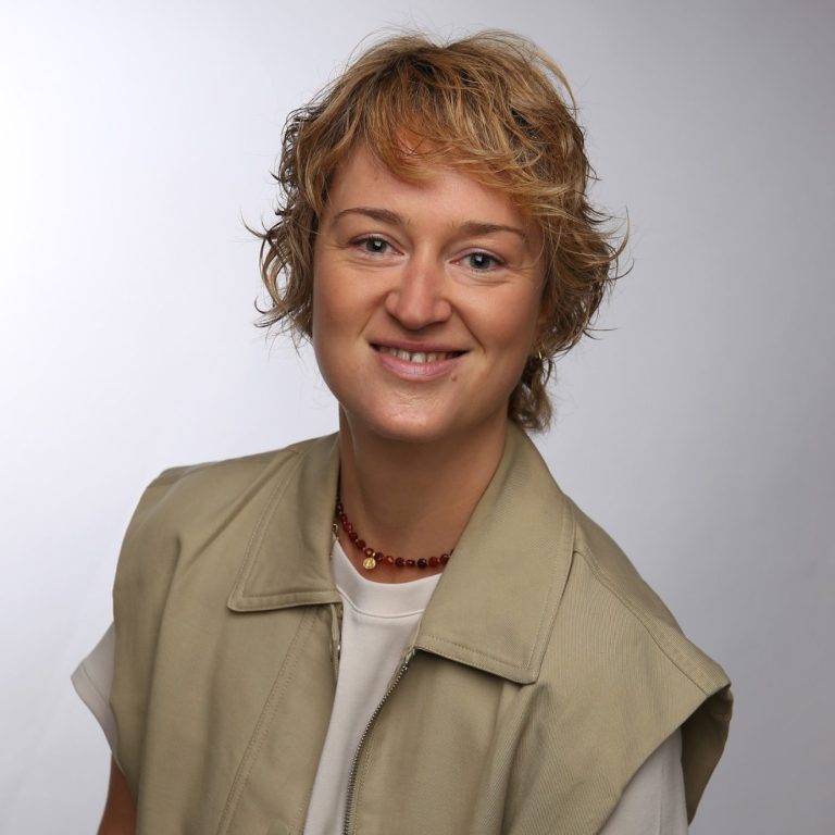 Felicitas Knapp – DiaVega – Deine vegane Ernährungsberatung Felicitas Knapp – eine weibliche Person mit kurzen, lockigen Haaren in beige Weste, lächelnd vor hellem Hintergrund.